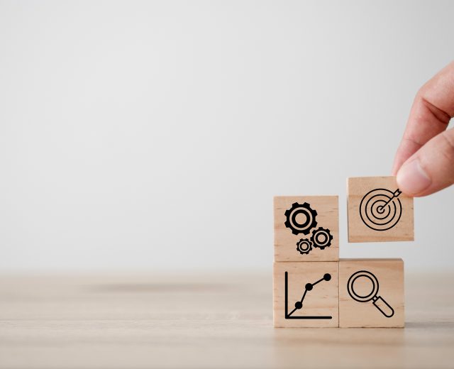 wooden blocks with gears, graph, target, and magnifying glass on them being stack into a square